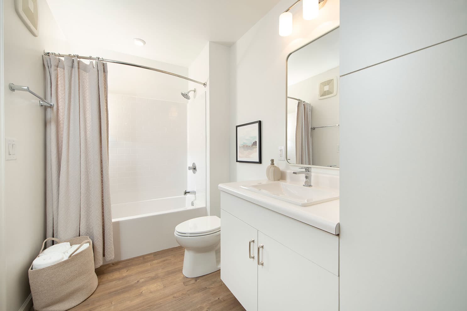 Bright bathroom with a white vanity, bathtub with a shower, and a basket of fresh towels on the wooden floor.