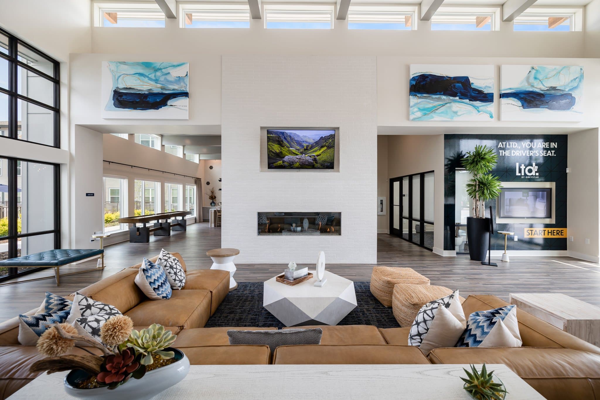Spacious and modern clubhouse interior with high ceilings, large abstract paintings, a central fireplace, and stylish lounge seating.