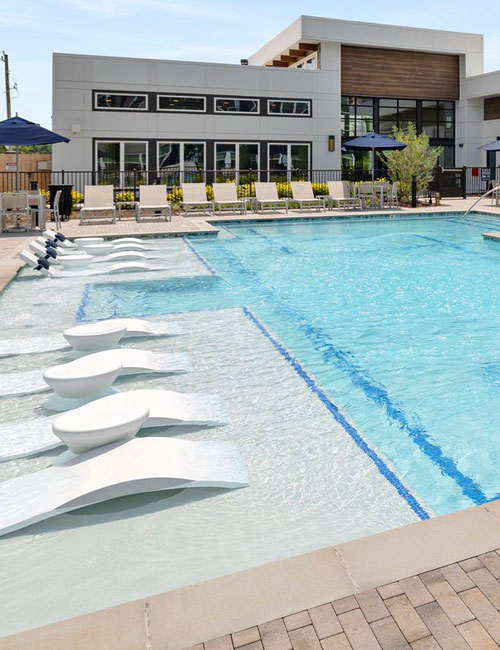 Contemporary swimming pool with in-water loungers, adjacent to a clubhouse with patio umbrellas and outdoor seating under a sunny sky.
